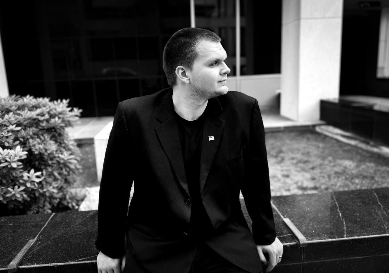 Isaac Mashman in a black blazer, seated on a stone ledge in downtown Little Rock, looking over his shoulder