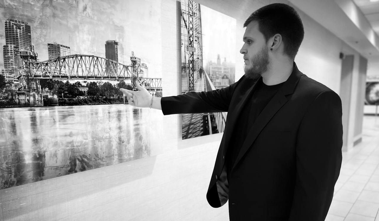 Isaac Mashman in a black blazer, pointing at a large canvas of the Little Rock skyline