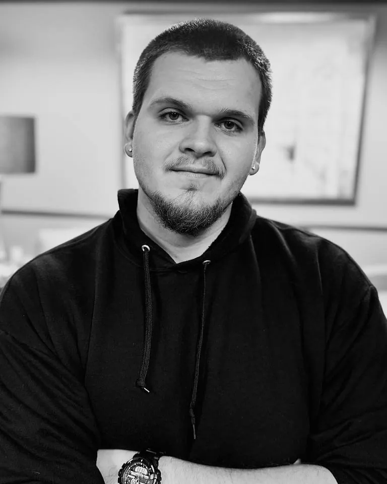 Isaac Mashman in a black hoodie, arms crossed, in a lounge with a lamp and white armchairs behind him