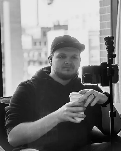 Isaac Mashman by a city window, a coffee in hand, beside a phone on a tripod