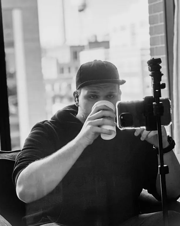 Isaac Mashman by a city window with a coffee, recording on a phone mounted on a tripod