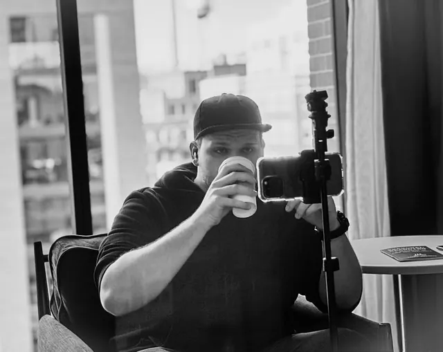 Isaac Mashman by a city window with a coffee, recording on a phone on a tripod