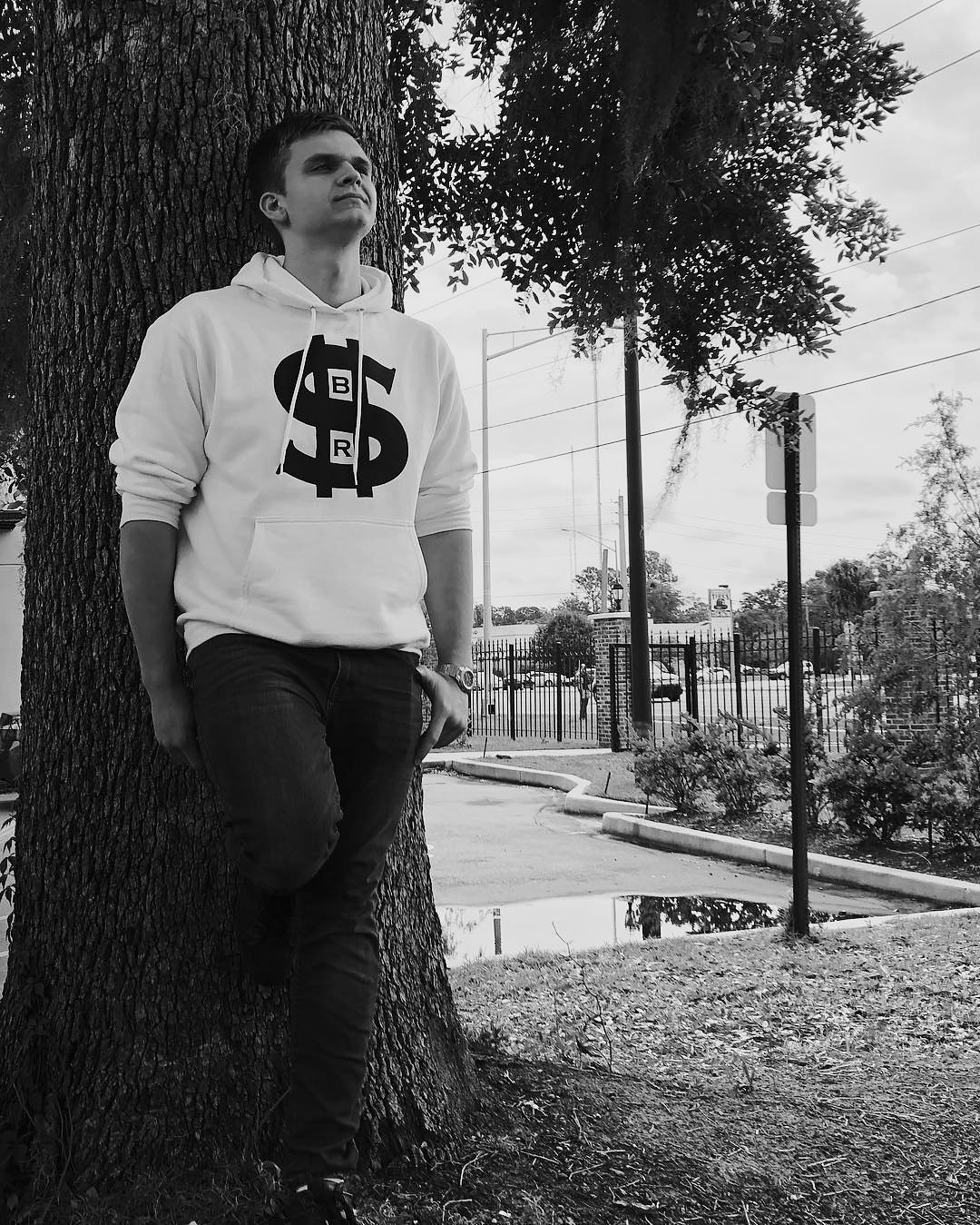 Isaac Mashman leaning against a tree in a white Success Before Rest Apparel hoodie with a dollar-sign logo